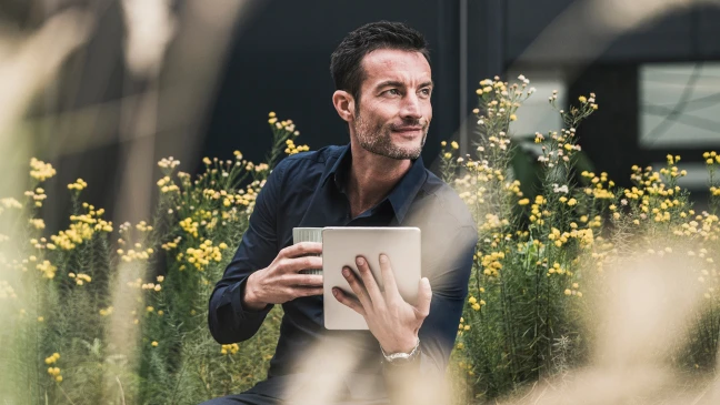 Person sitzt im Freien inmitten gelber Blüten und hält ein Tablet und einen Becher, während im Hintergrund dunkle Gebäudeteile sichtbar sind.