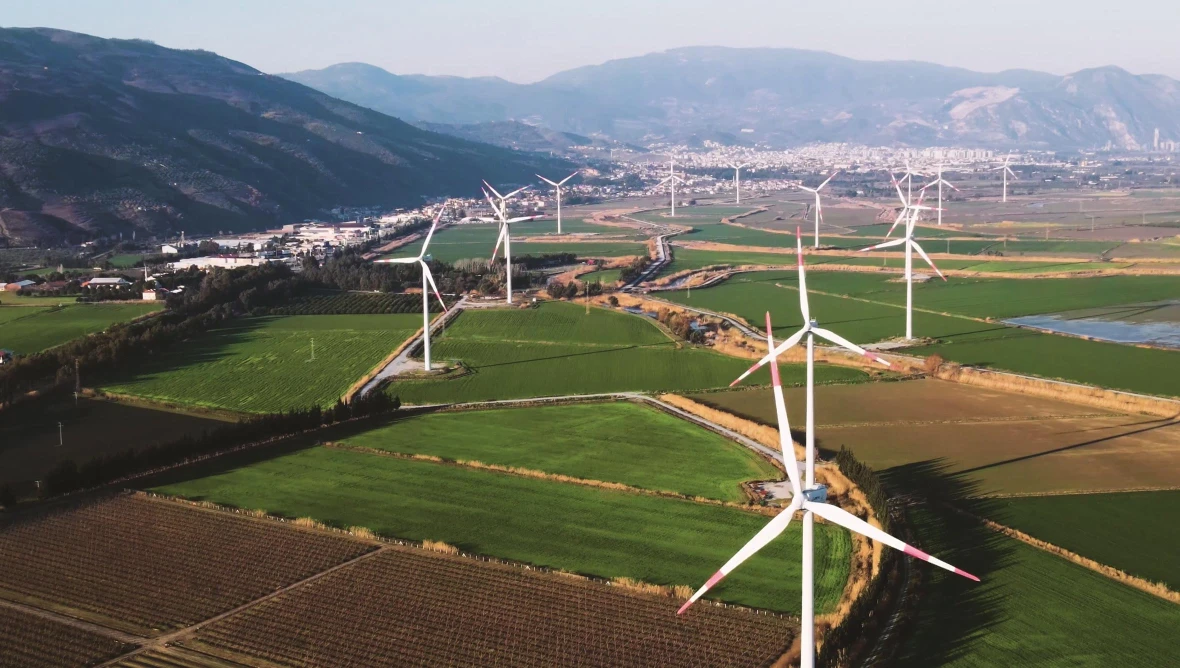 Windräder stehen auf grünen Feldern vor einer Bergkulisse, im Hintergrund ist eine Stadt erkennbar.