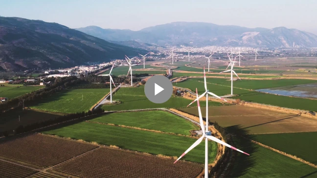 Windräder stehen auf grünen Feldern vor einer Bergkulisse, im Hintergrund ist eine Stadt erkennbar.