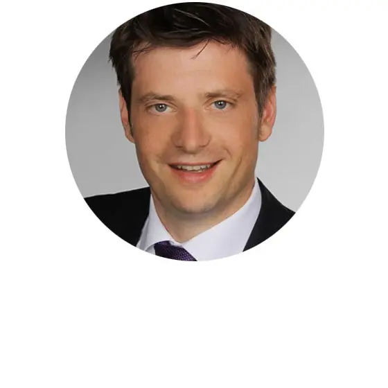 Eine Person mit kurzen braunen Haaren in einem formellen Anzug und einer lila Krawatte lächelt. Der Hintergrund ist unscharf.