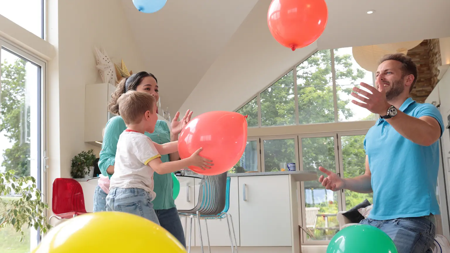 Person mit langen Haaren und rotem Oberteil sitzt an einem Tisch und schaut lächelnd auf einen Laptop vor einem Fenster.