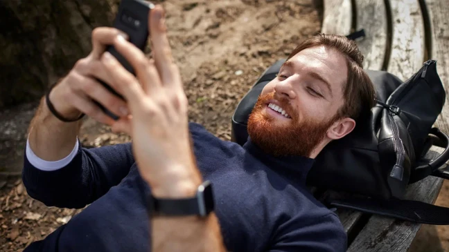 Person mit rötlich-braunem Bart liegt auf Holzbank, hält Smartphone in der rechten Hand und lächelt beim Blick auf den Bildschirm.