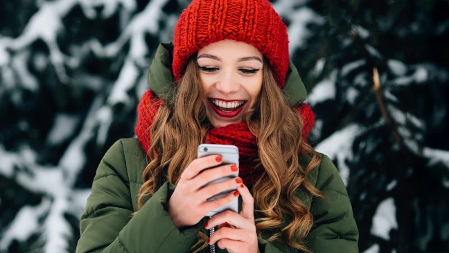 Frau mit roter Mütze steht vor verschneiten Tannen und hält Handy in der Hand