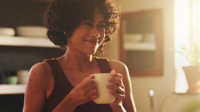 Person hält eine große Tasse in beiden Händen und steht in einem warm beleuchteten Raum; im Hintergrund sind Küchenregale und eine Fensterreflexion sichtbar.