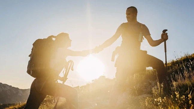 Zwei Personen mit Wanderausrüstung befinden sich an einem steilen Berghang im Gegenlicht der Sonne. Eine Person kniet leicht erhöht und hält einen Wanderstock, während sie der anderen Person die Hand reicht; im Hintergrund sind Berge, Wiesen und ein klarer Himmel zu sehen.