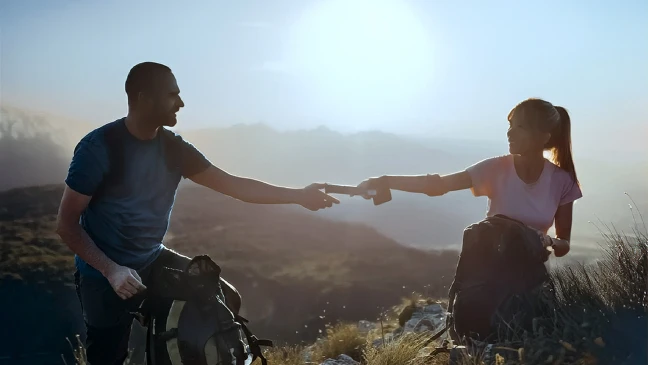 Zwei Personen mit Rucksäcken reichen sich auf einem Berggipfel einen Gegenstand vor einer Landschaft im Abendlicht.