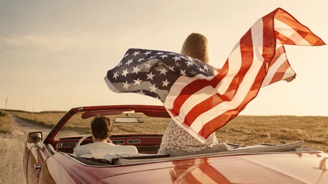 Person mit US-Flagge im Cabrio auf staubiger Landstraße bei Sonnenuntergang