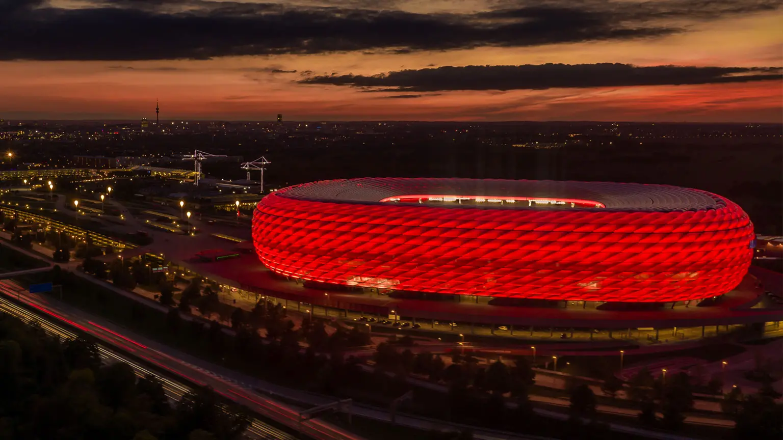 Rotes Trikot des FC Bayern München mit Sponsorenlogos, präsentiert vor der Allianz Arena bei Sonnenuntergang davor ein rot-weißer Fußball mit Vereinslogo.