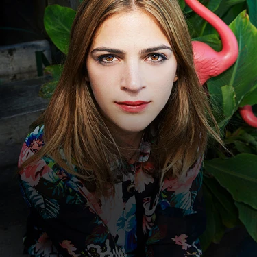 Person mit blonden Haaren vor tropischen Pflanzen und zwei rosa Plastikflamingos, trägt bunte Bluse, schaut direkt in die Kamera., mit Allianzlogo.