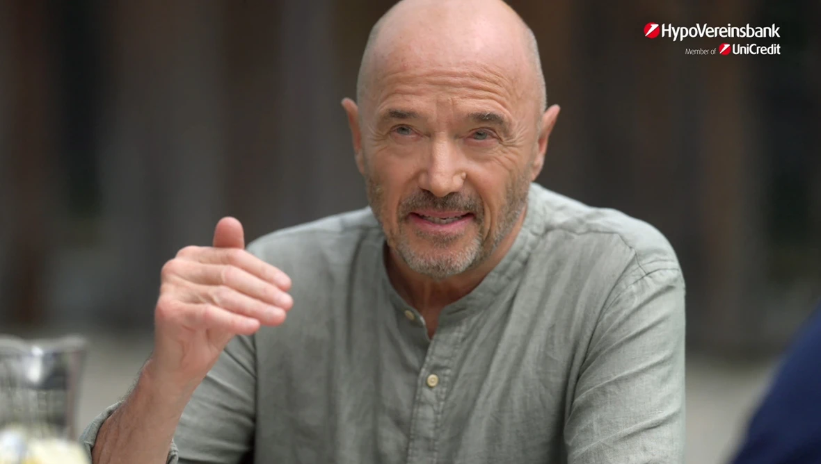 Christian Neureuther in grauem Hemd mit erhobener Hand, Glasbehälter mit Wasser links, Logo HypoVereinsbank oben rechts.