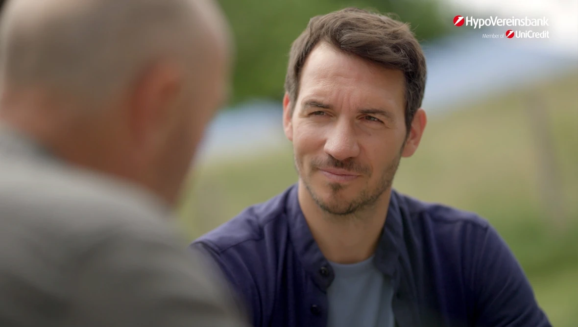 Felix und Christian Neureuther sitzen draußen an einem Tisch, im Hintergrund grüne Bäume und ein Zaun, oben rechts das HypoVereinsbank Logo.