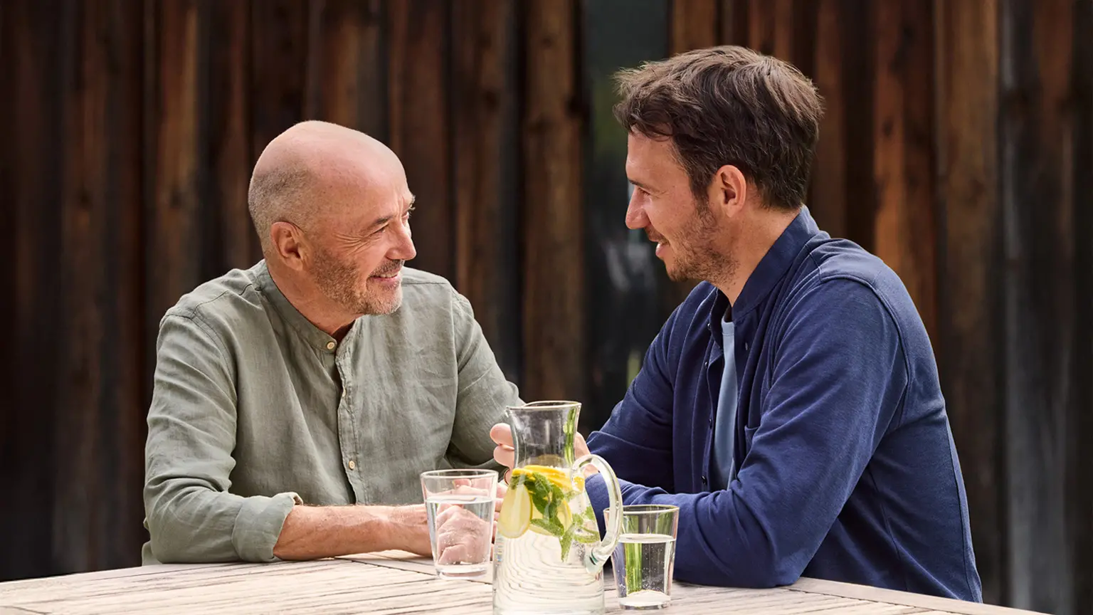 Christian und Felix Neureuther sitzen an einem Holztisch mit Wasserkrug und Gläsern, im Hintergrund dunkle Holzfläche.