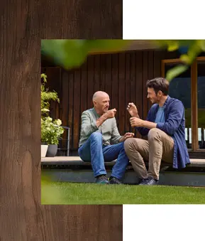 Christian und Felix Neureuther sitzen auf Holzterrasse vor dunkler Holzfassade, grüne Pflanzen links, Rasen im Vordergrund, Fenster im Hintergrund.