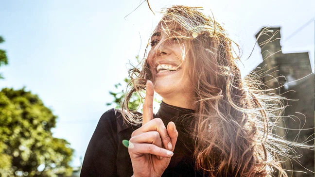 Lachende Frau mit wehenden Haaren zeigt mit dem Finger nach oben, im Hintergrund Bäume und ein Gebäude bei Sonnenschein.
