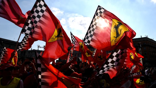 Mehrere rote Ferrari-Flaggen mit schwarzem Pferde-Logo und schwarz-weiß kariertem Rand wehen im Wind vor blauem Himmel. 