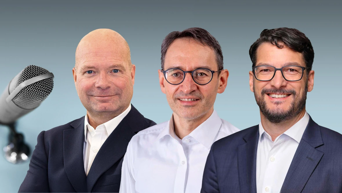 Drei Männer in formeller Kleidung stehen nebeneinander. Links ein glatzköpfiger Mann im dunklen Anzug mit weißem Hemd, in der Mitte ein Mann mit kurzen dunklen Haaren, Brille und hellem Hemd, rechts ein Mann mit kurzen dunklen Haaren, Bart, Brille und dunklem Anzug mit weißem Hemd. Links im Bild ist ein Mikrofon sichtbar.