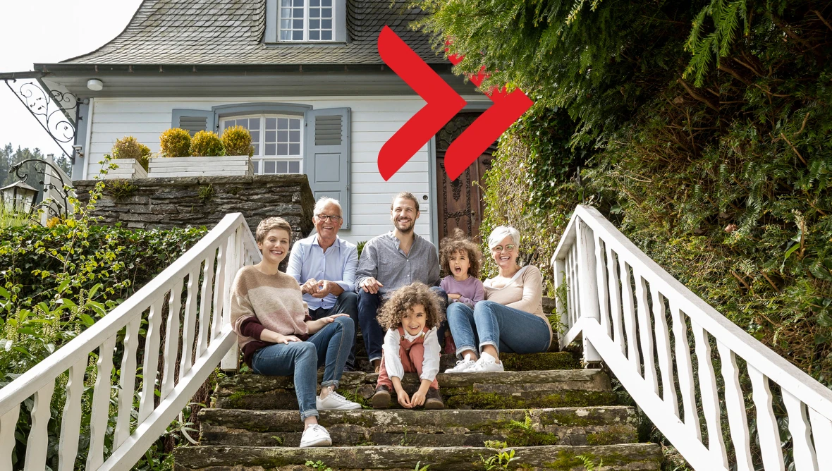 Mehrere Personen sitzen gemeinsam auf einer steinernen Außentreppe vor einem hellen Haus mit Fensterläden und Gartenpflanzen. Im Vordergrund sitzen zwei Kinder, dahinter weitere Personen. Weiße Geländer, begrünte Umgebung und Tageslicht prägen die Szene.
