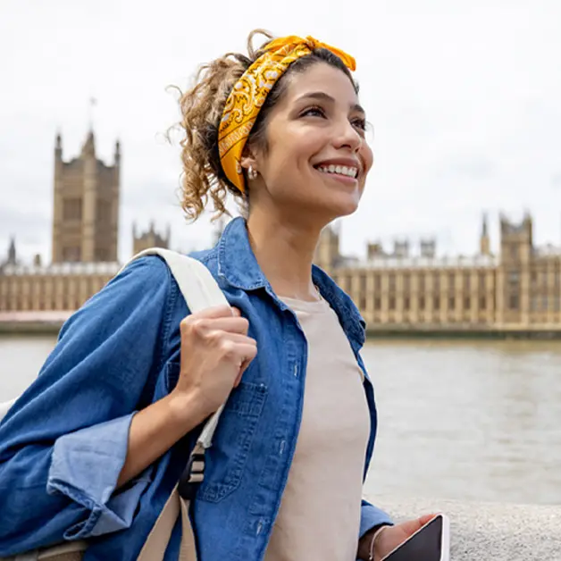 Person mit gelbem Kopftuch und Jeanshemd steht vor historischem Gebäude an Fluss, rechts ein Turm mit Uhr.