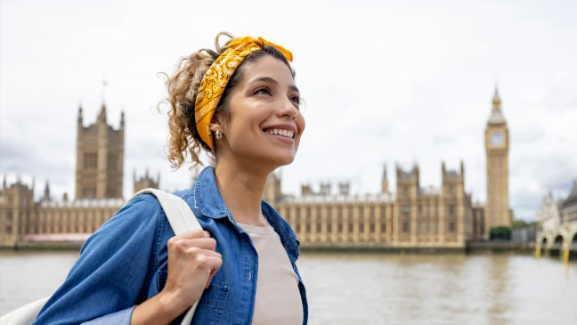 Eine Person mit Rucksack steht an einem Flussufer vor großen historischen Gebäuden und einem hohen Turm mit Uhr, der deutlich im Hintergrund sichtbar ist.