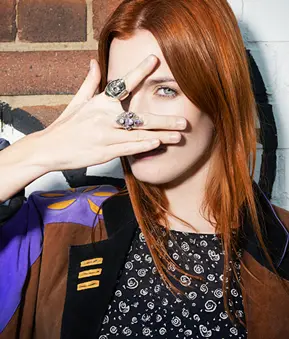 Person mit goldenem Gesichtsmake-up und schwarzem Kleidungsstück blickt in die Kamera. Hintergrund ist unscharf.