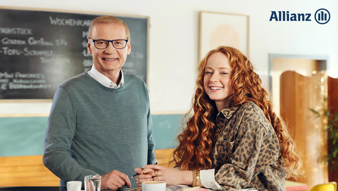 Günther Jauch und weitere Person im Café am Tisch, im Hintergrund Menütafel und Allianz-Logo sichtbar.