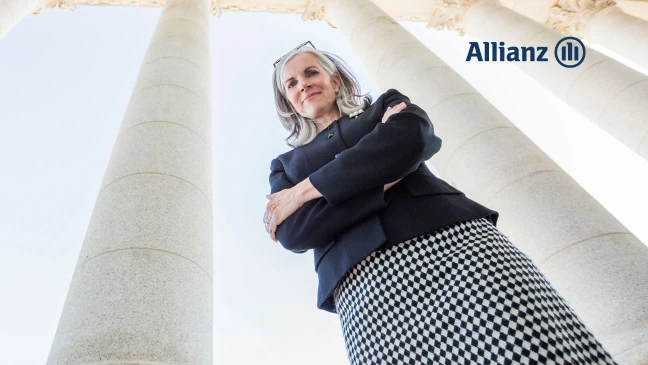 Person in Business‑Outfit steht mit verschränkten Armen vor hohen Steinsäulen, aus einer niedrigen Perspektive fotografiert; rechts oben ist das Allianz‑Logo zu sehen.