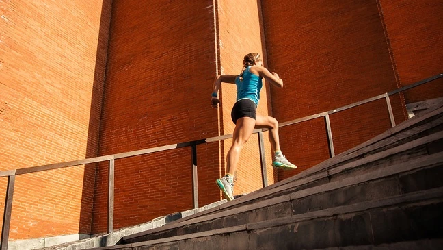 Eine Person läuft eine breite Treppe hinauf, die vor einer hohen Backsteinwand liegt, während Sonnenlicht von der Seite auf die Szene fällt.