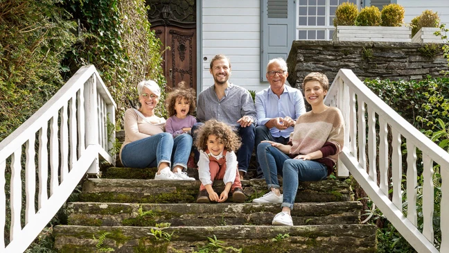 Mehrere Personen sitzen auf einer Außentreppe vor einem Wohnhaus; Kinder und Erwachsene gemeinsam im Grünen.