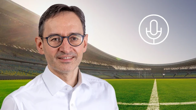 Person mit Brille und weißem Hemd vor Fußballstadion, rechts ein Mikrofon‑Symbol, Spielfeld und Tribünen im Hintergrund.