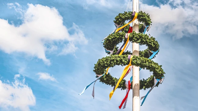 Maibaum mit grünem Kranzschmuck und bunten Bändern vor blauem Himmel, Symbol für Frühling und Tradition.
