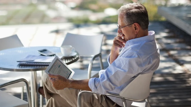 Person sitzt an einem runden Tisch im Freien und hält ein Tablet. Auf dem Tisch liegen ein Notizbuch, ein Stift und eine weiße Tasse.