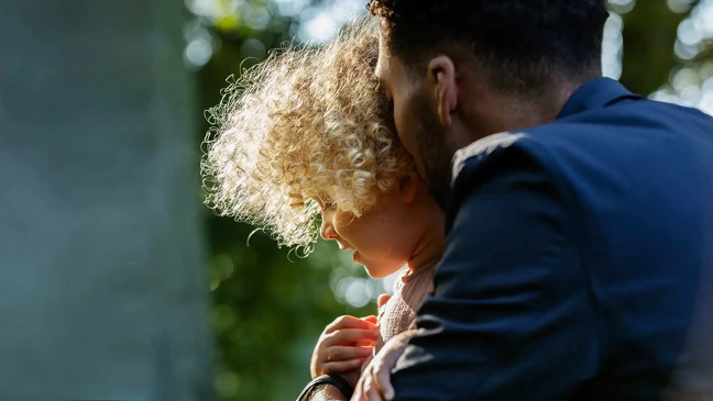 Erwachsene Person hält ein kleines Kind mit lockigem Haar im Arm im Freien mit unscharfem grünem Hintergrund und seitlichem Sonnenlicht.