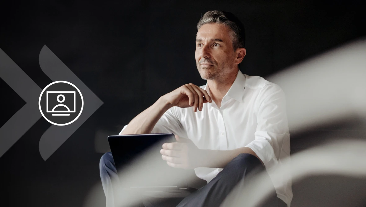 Person in weißem Hemd sitzt mit Laptop in nachdenklicher Haltung vor dunklem Hintergrund.