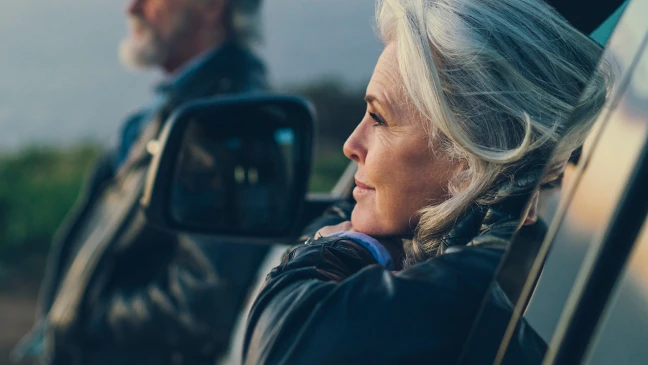 Eine Person lehnt an einer offenen Autotür, während eine weitere Person im Hintergrund neben dem Fahrzeug steht, umgeben von Natur.