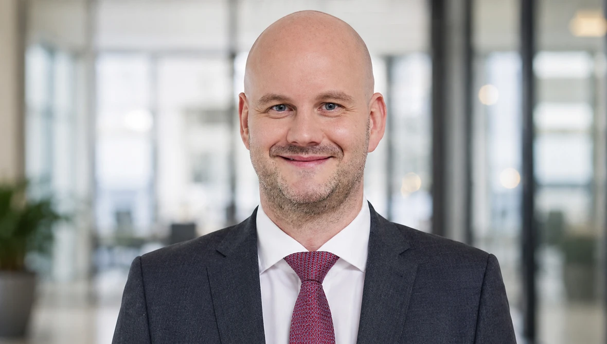 Portrait einer Person in dunklem Anzug und weißem Hemd mit roter Krawatte vor unscharfem Bürohintergrund.