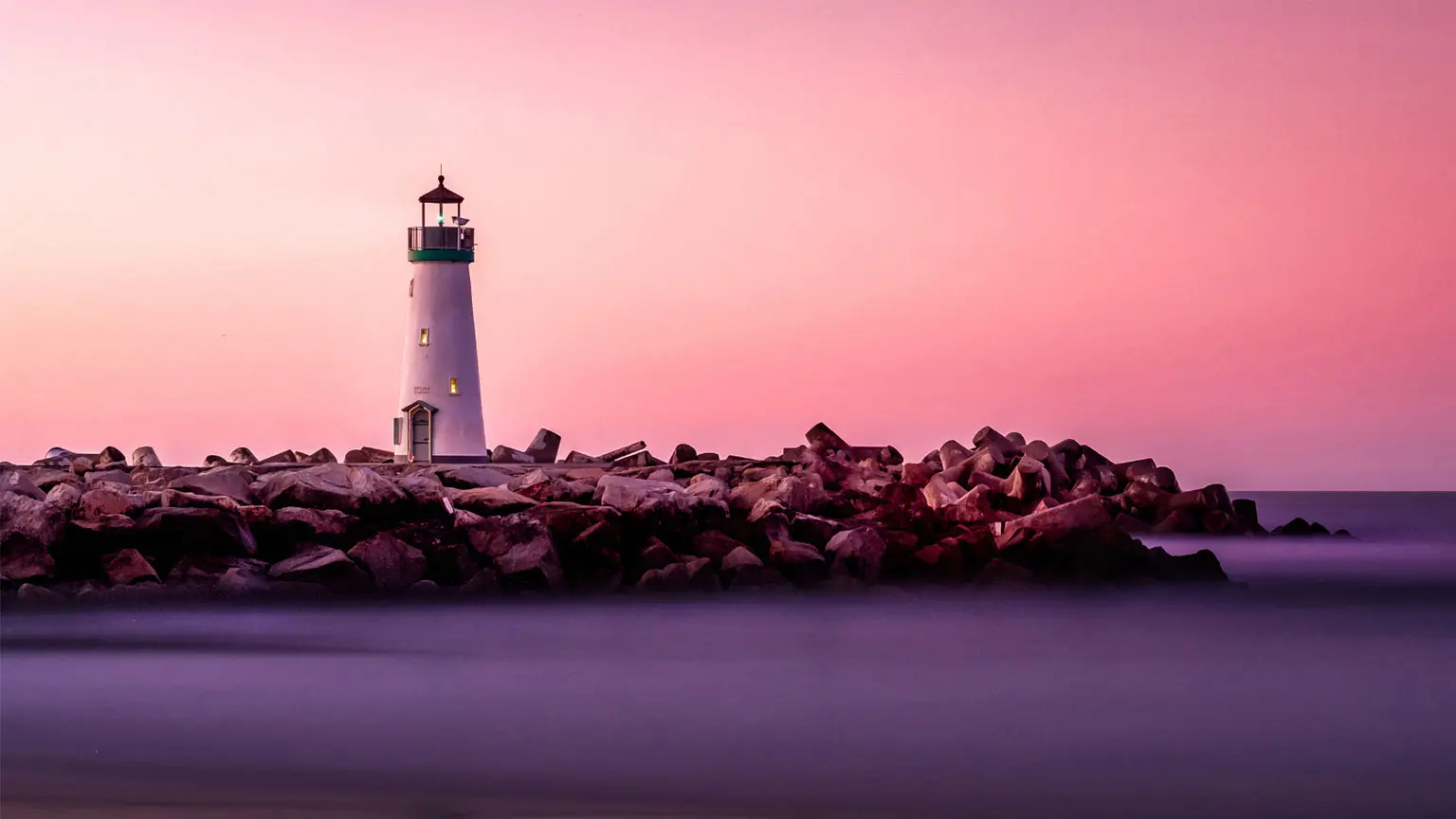 A white lighthouse stands on rocks by the water against a pink-purple sky.