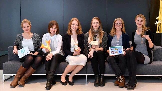 Sechs Frauen sitzen auf einem Sofa, halten verschiedene Gegenstände in den Händen und blicken lächelnd in die Kamera vor dunklem Hintergrund.