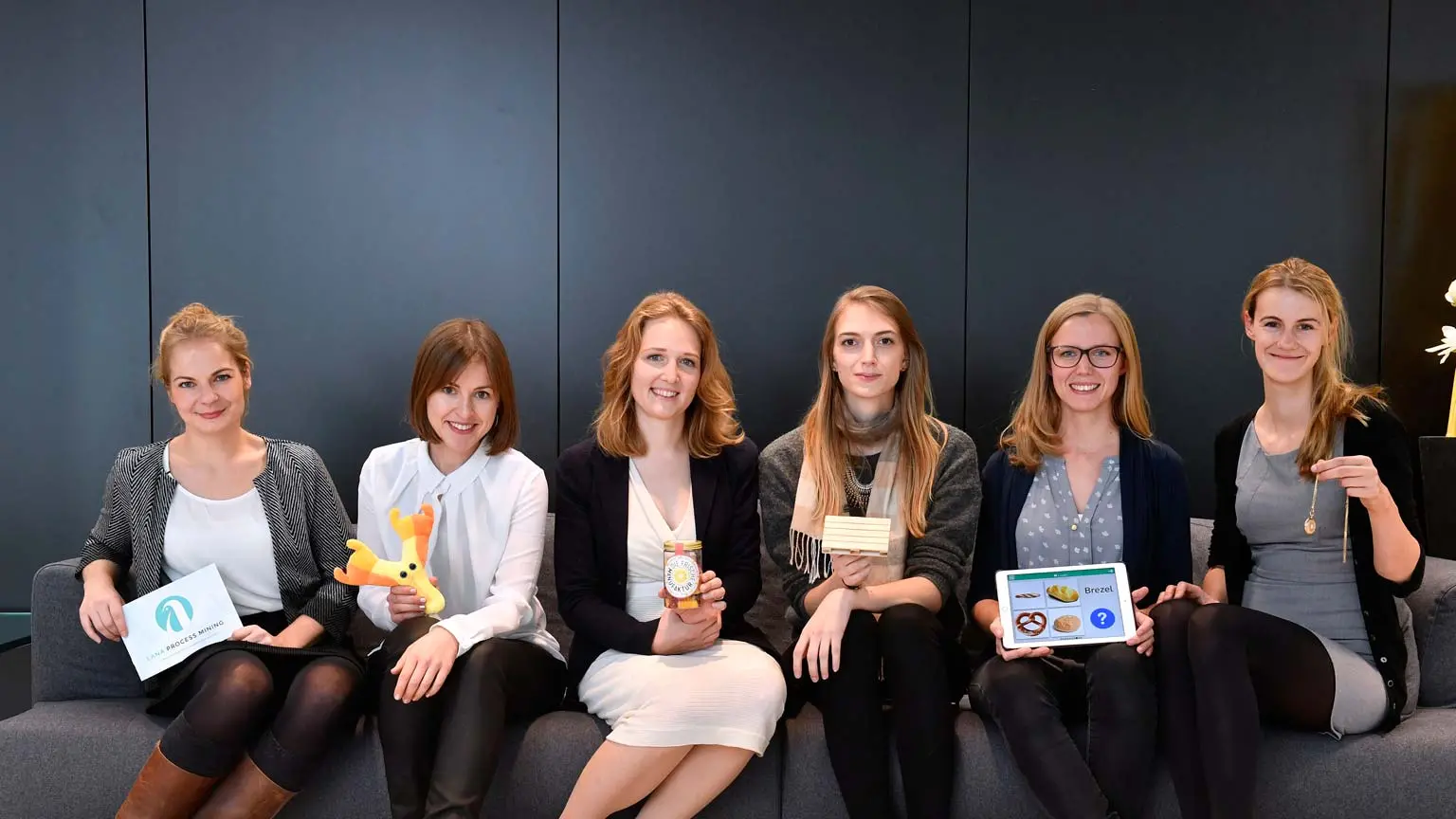 Sechs Frauen sitzen auf einem Sofa, halten verschiedene Gegenstände in den Händen und blicken lächelnd in die Kamera vor dunklem Hintergrund.