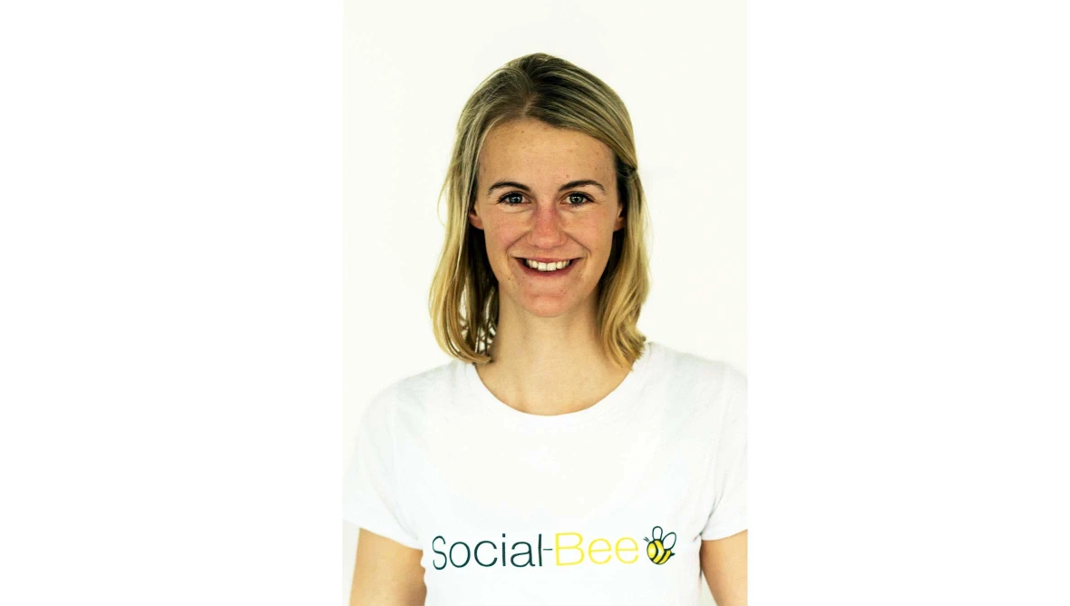 Lächelnde Frau mit mittellangem blondem Haar trägt ein weißes T-Shirt mit der Aufschrift „Social-Bee“ und einem stilisierten Bienenlogo.