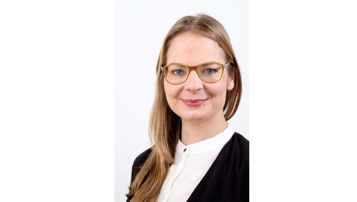 Frau mit langen Haaren, goldfarbener Brille, weißer Bluse und schwarzem Blazer vor hellem Hintergrund, schaut freundlich in die Kamera.