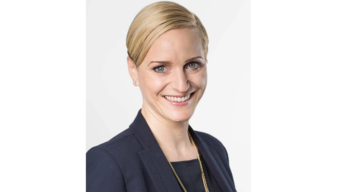 Frau mit blondem, zurückgekämmtem Haar trägt dunkles Sakko, dunkles Oberteil und goldene Halskette vor weißem Hintergrund.