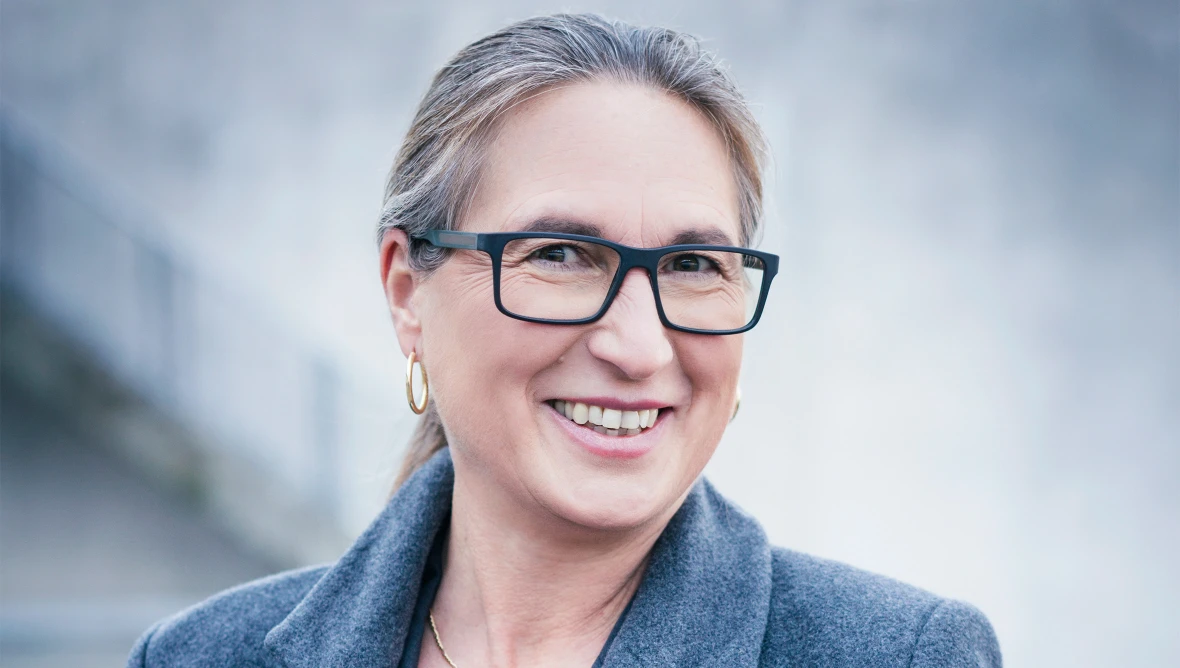 Close-up of Marion Höllinger, smiling, wearing glasses, with gray hair and a dark gray coat, against a blurred, bright outdoor background.