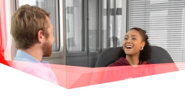 Two people indoors in conversation; one with light hair facing away, the other with dark curly hair, smiling; window blinds in background.