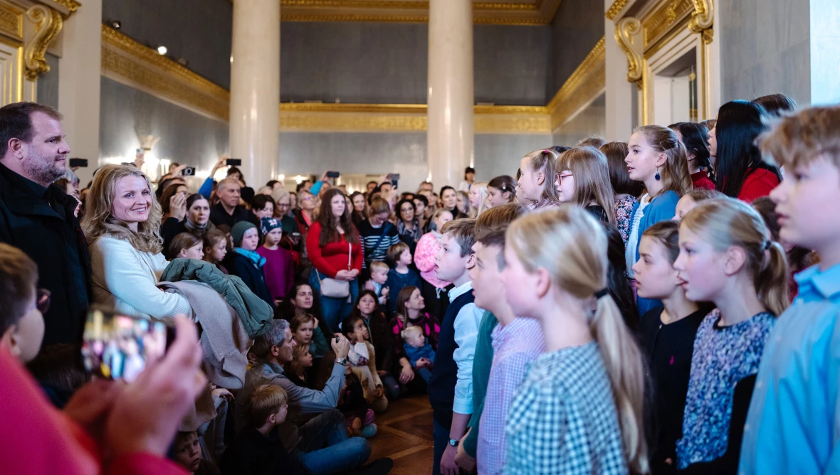 Auf der rechten Bildseite stehen Kinder hintereinander zu einem Chor aufgestellt, links stehen und sitzen dicht gedrängt Erwachsene und Kinder und hören dem Chor zu.