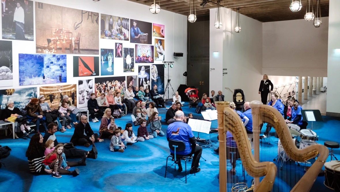 Großer Raum mit blauem Teppich und großen Bildern an der Wand, in der Mitte sitzen Musiker:innen in blauen T-Shirts, davor sitzen einige Kinder und Eltern auf dem Fußboden und schauen gespannt den Musiker:innen zu.