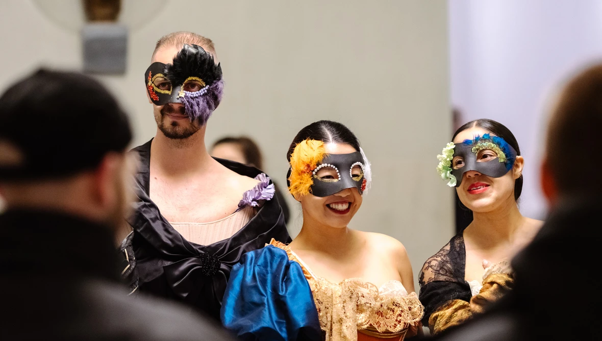 Ein Mann und zwei Frauen sind verkleidet mit Masken im Gesicht und bunten Gewändern, man sieht alle drei vom Kopf bis zum Oberkörper