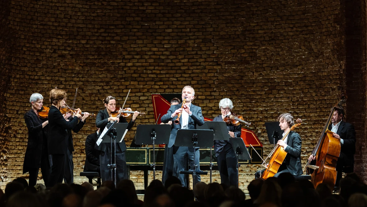 Neun Personen stehen auf einer Bühne, vier Frauen spielen Geige, ein weiterer Herr spielt Geige, ein Mann Kontrbass und ein Mann Cello. Im Vordergrund spielt ein Mann Klarinette und ganz im Hintergrund sitzt ein Mann am Flügel.