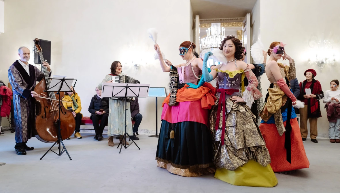 Drei Personen in bunten langen Gewändern tanzen auf der rechten Seite des Fotos, links steht ein Musiker mit einem Kontrabass und eine Musikerin mit einer Ziehharmonika.