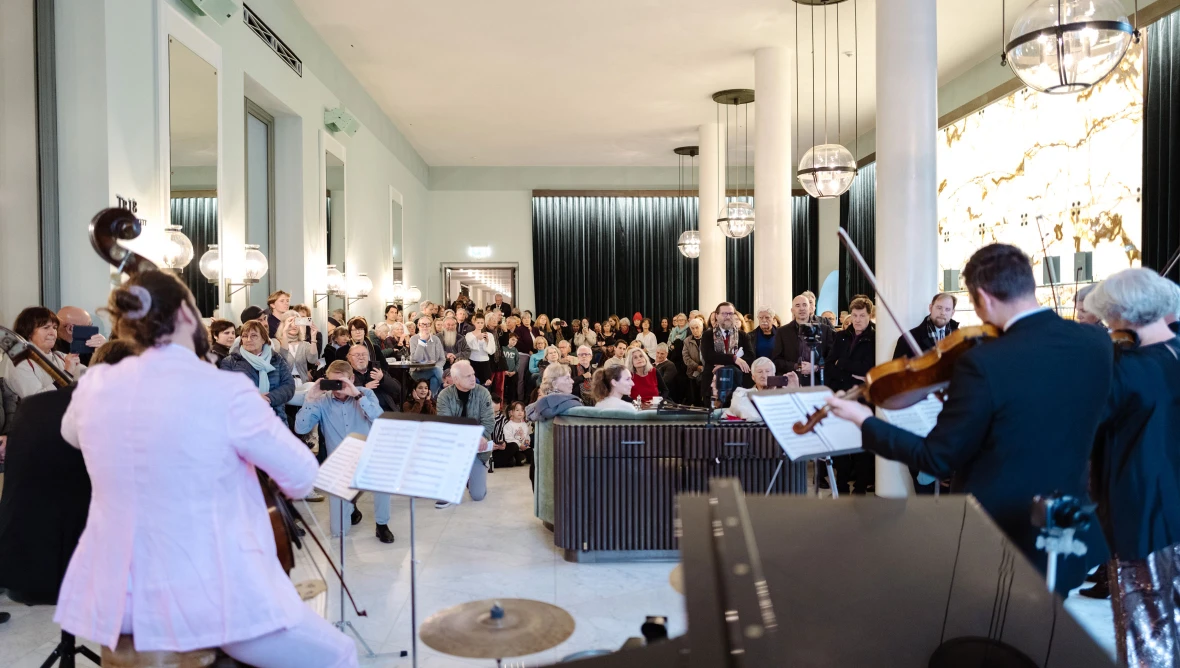 Blick von hinten auf Musiker:innen, die Geige und Kontrabass spielen, im Hintergrund sieht man die Räumlichkeiten einer Bar, darin sitzen und stehen zahlreiche Menschen, die den Musiker:innen zusehen und zuhören.