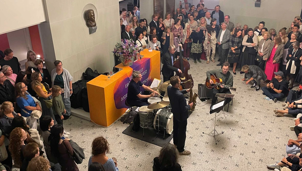 Viele Personen umgeben eine Musikgruppe mit Schlagzeug Kontrabass Gitarre und Saxofon in einem Innenraum.